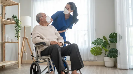 Portrait of Asian caregiver nurse take care senior male on wheelchair. Attractive specialist carer women wear face mask, support elderly grandfather at nursing home care and smile, look at each other