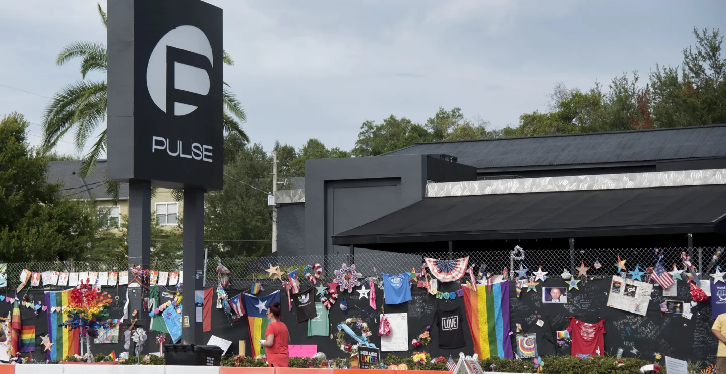 A sign for the Pulse nightclub on the left with flowers, shirts, rainbow flags and other memorial items hung on a chainlink fence to the right in memory of those killed in a 2016 mass shooting.