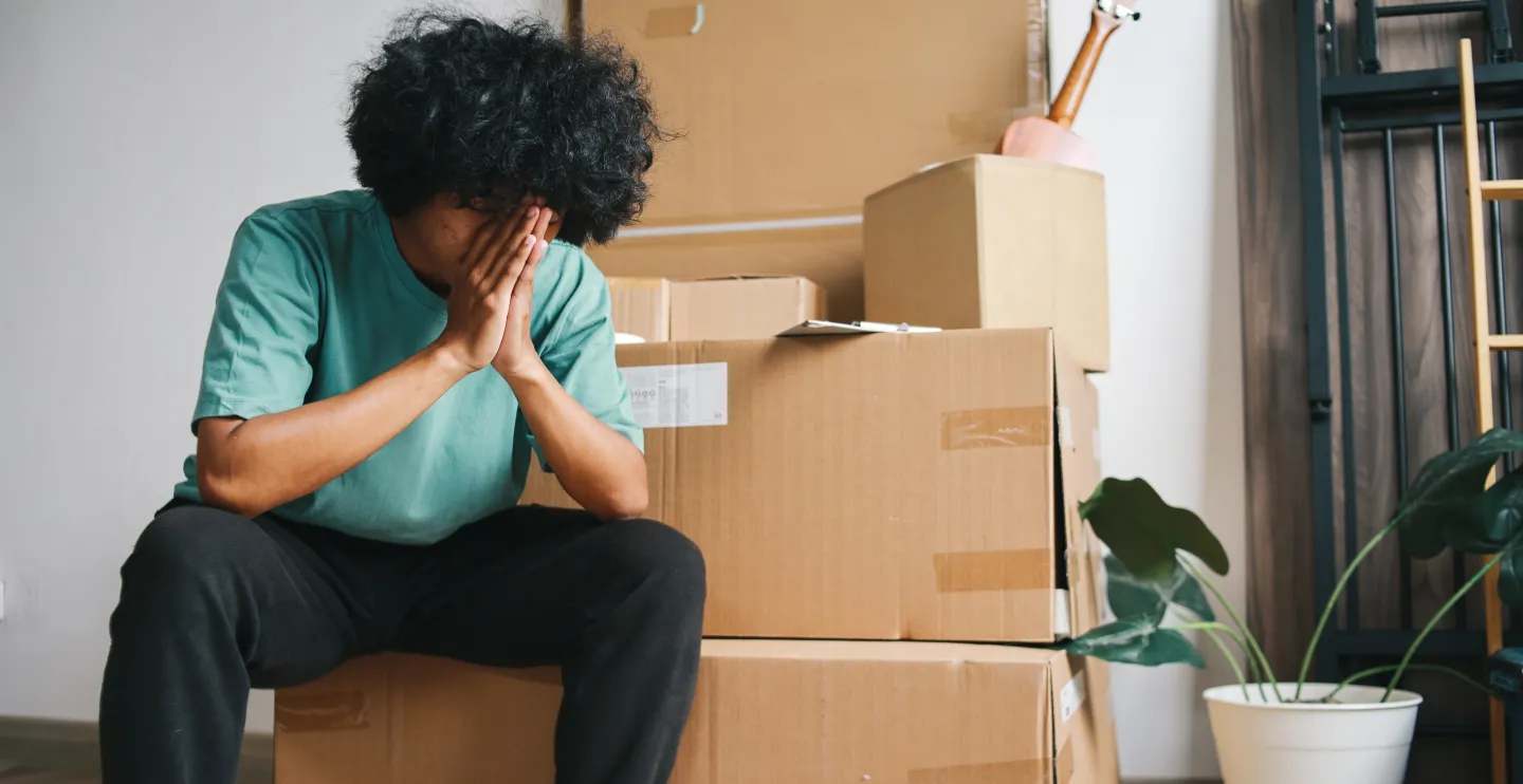 Man with dark curly hair sitting with his face in his hands in front of a pile of cardboard boxes as if stressed out about moving.