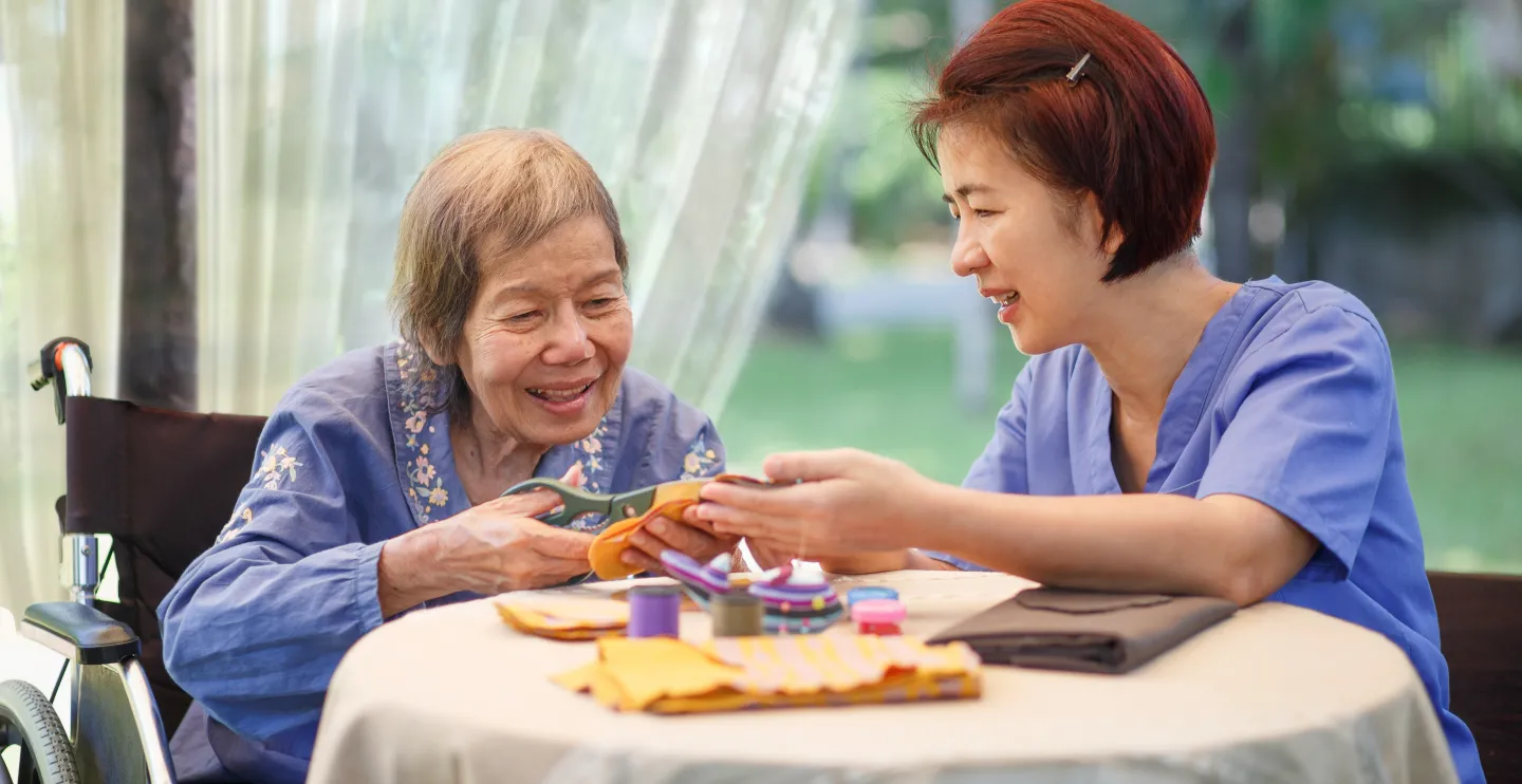 Older Asian woman at left with Asian woman caregiver who is helping her make crafts as they both sit at a table.