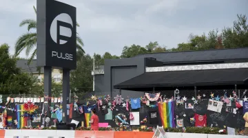 A sign for the Pulse nightclub on the left with flowers, shirts, rainbow flags and other memorial items hung on a chainlink fence to the right in memory of those killed in a 2016 mass shooting.