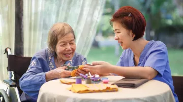 older woman using scissors with the support of an aide wearing scrubs