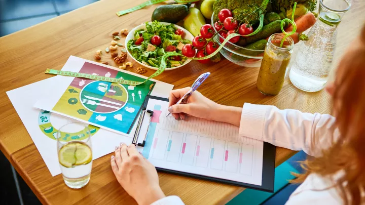 Woman dietitian in medical uniform with tape measure working on a diet plan sitting with different healthy food ingredients in the green office on background.